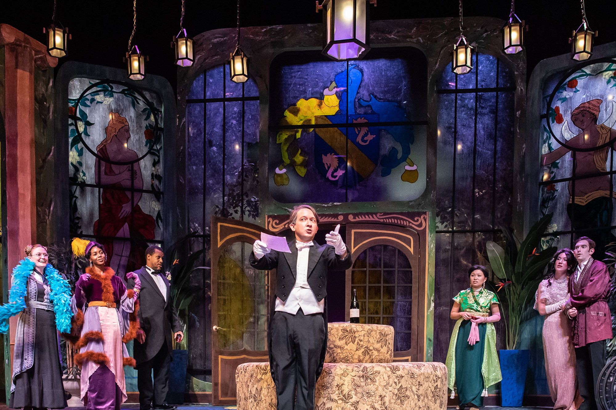 Scene during a play in which a group of well-dressed people surround a man in a tuxedo and listen with curiosity