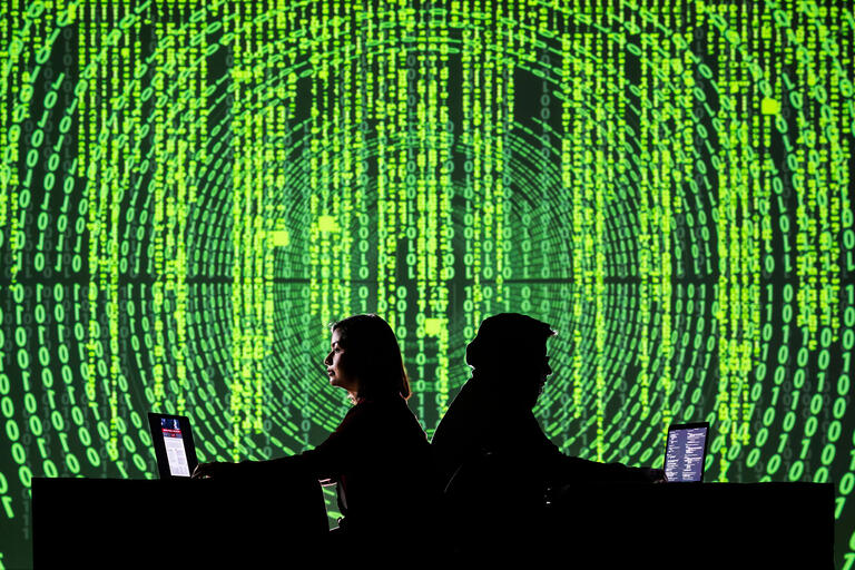 two people back to back at computer screens with code projected behind them