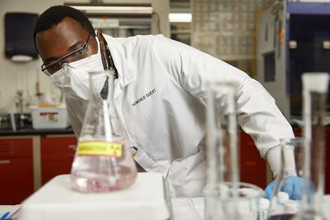 Person in lab coat and mask observing a beaker